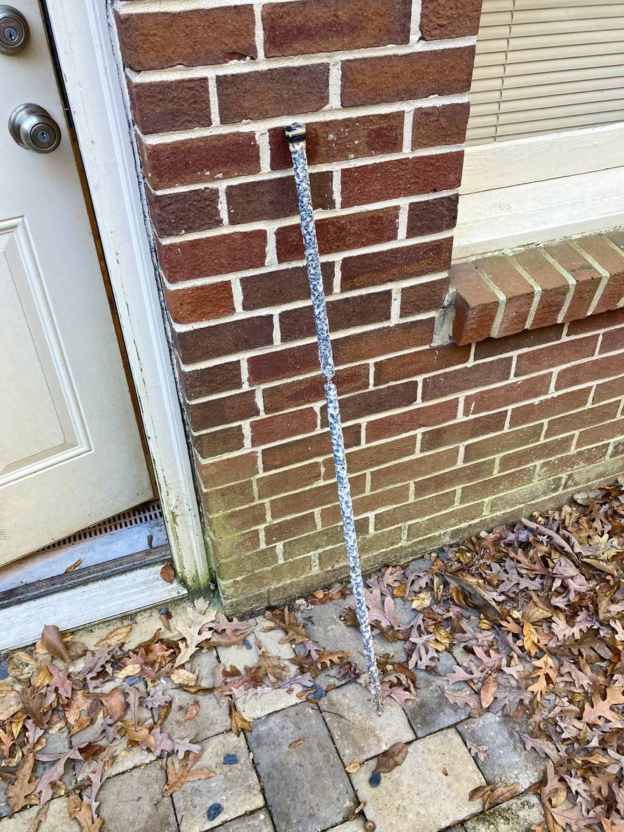 A long corroded rod leaning against the exterior of a brick house.