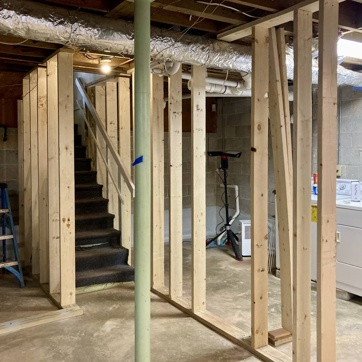 An unfinished basement with studded walls up. A green pole is in the middle and some stairs behind it.