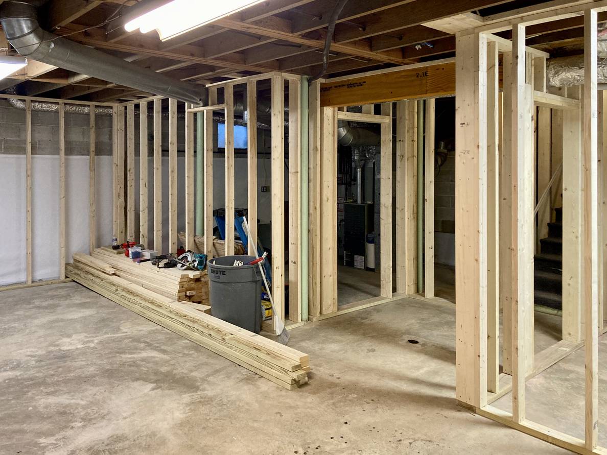 An unfinished basement with rooms delineated by wood stud walls. A large wooden header spans an opening at the foot of the stairs.