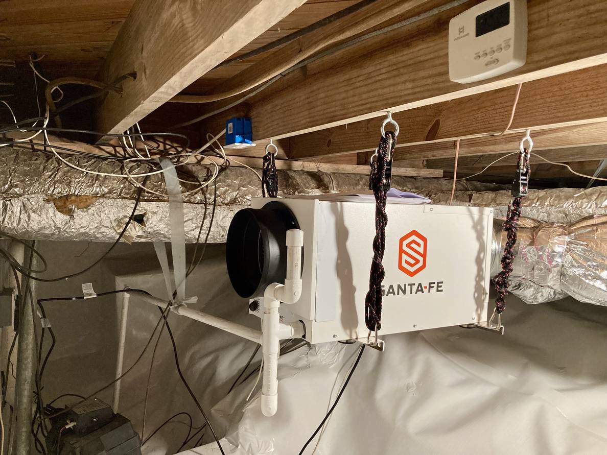 A boxy metal dehumidifier hangs from floor joists in a crawlspace. The ground is covered in white plastic. Wire, ducting, and pvc pipes are throughout the image.