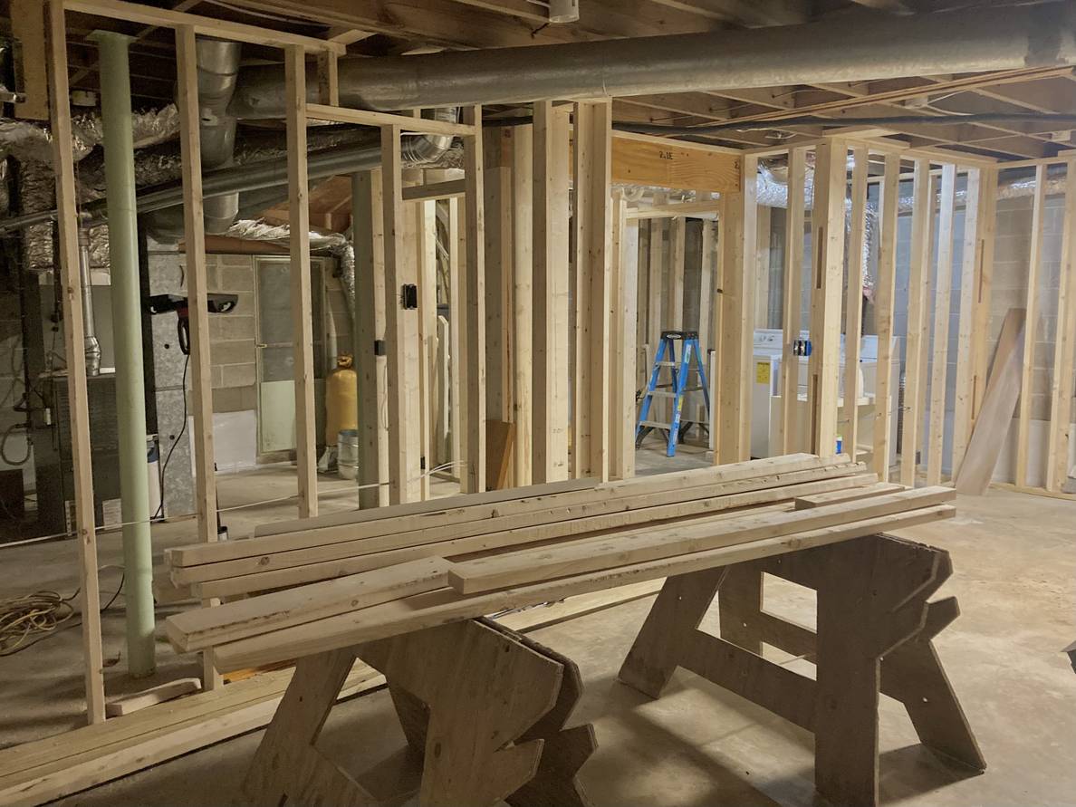 A basement with framed walls. In the foreground is a couple of sawhorses holding lumber. Behind the framed walls on the left is a space with an HVAC unit. The framed walls open into an alcove.