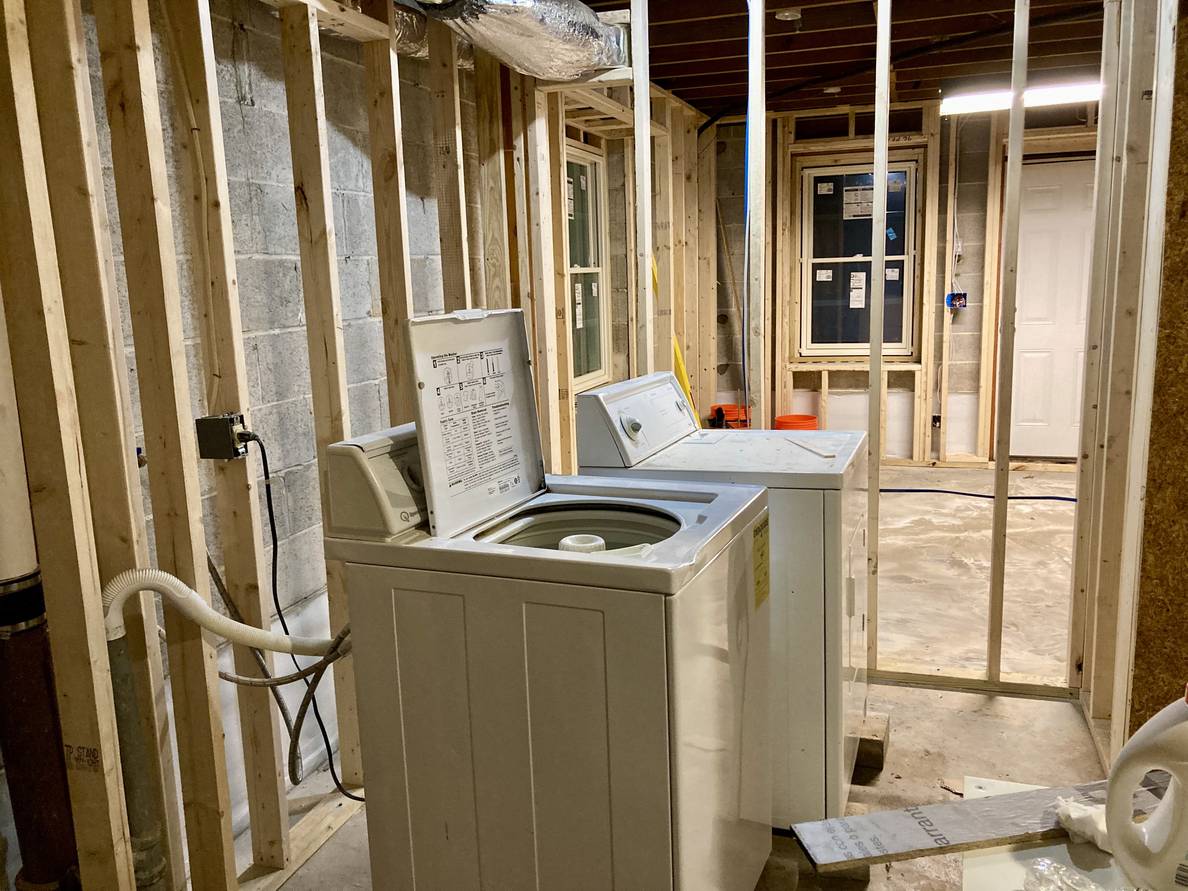 The same unfinished laundry room from the opposite wall. The laundry machines are still in frame, and through the wall beyond them is the main basement space.