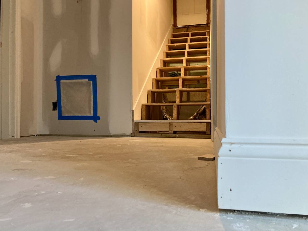 A photo a partially finished basement. The angle is close to the ground and close to the camera is a wall corner with unpainted baseboard. In the background is a flight of unfinished stairs with some trim installed on the wall along the steps.