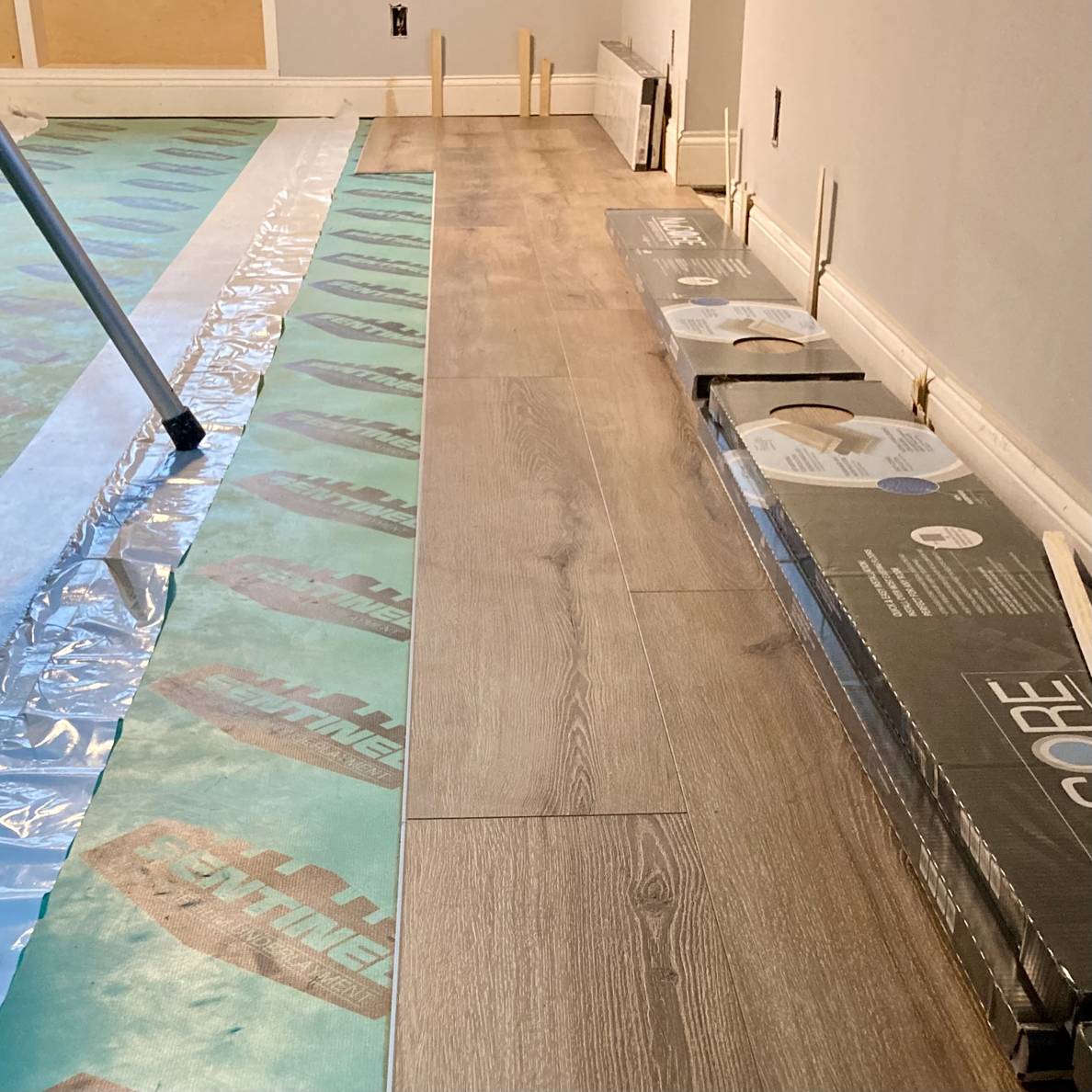 A photo of a partially finished basement floor. Most of the floor is covered by underlayment, a few rows of wood-looking flooring has been installed.