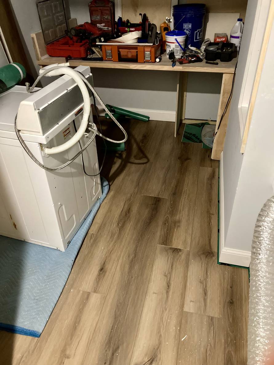 A photo of a partially finished laundry room with flooring installed. The laundry machines are moved away from the wall and a smattering of tools are on a shelf.