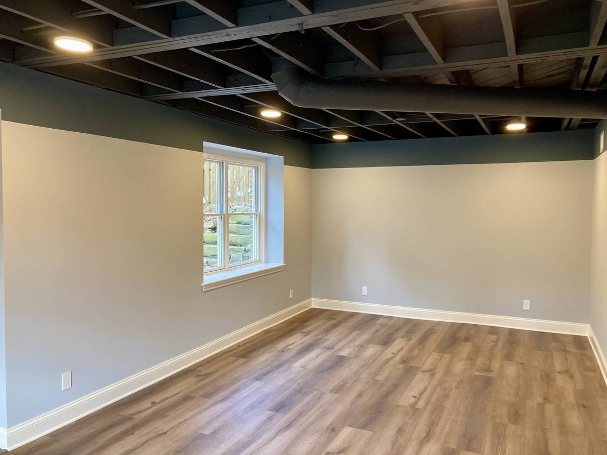 A practically finished basement living space. The ceiling is painted a charcoal gray with circular lights. A bluish gray foot-long-ish border goes along the top of the walls. The bulk of the walls are a gray color. The window well is inset against the border. The flooring is a wood-looking material.