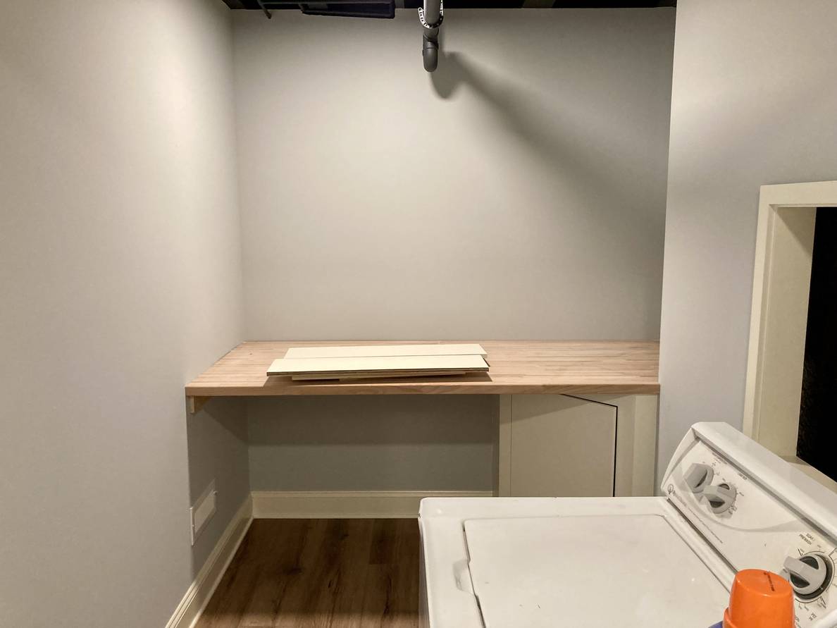 A photo of the laundry room. A wooden laundry shelf spans the far wall. The wall to the right has a framed opening where the the plumbing can be accessed.