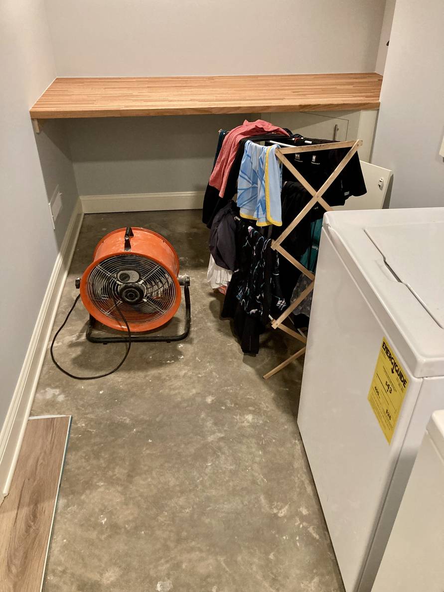 A photo of the bare concrete floor of the laundry room where flooring used to be. A single plank of flooring is still down towards the door of the room.