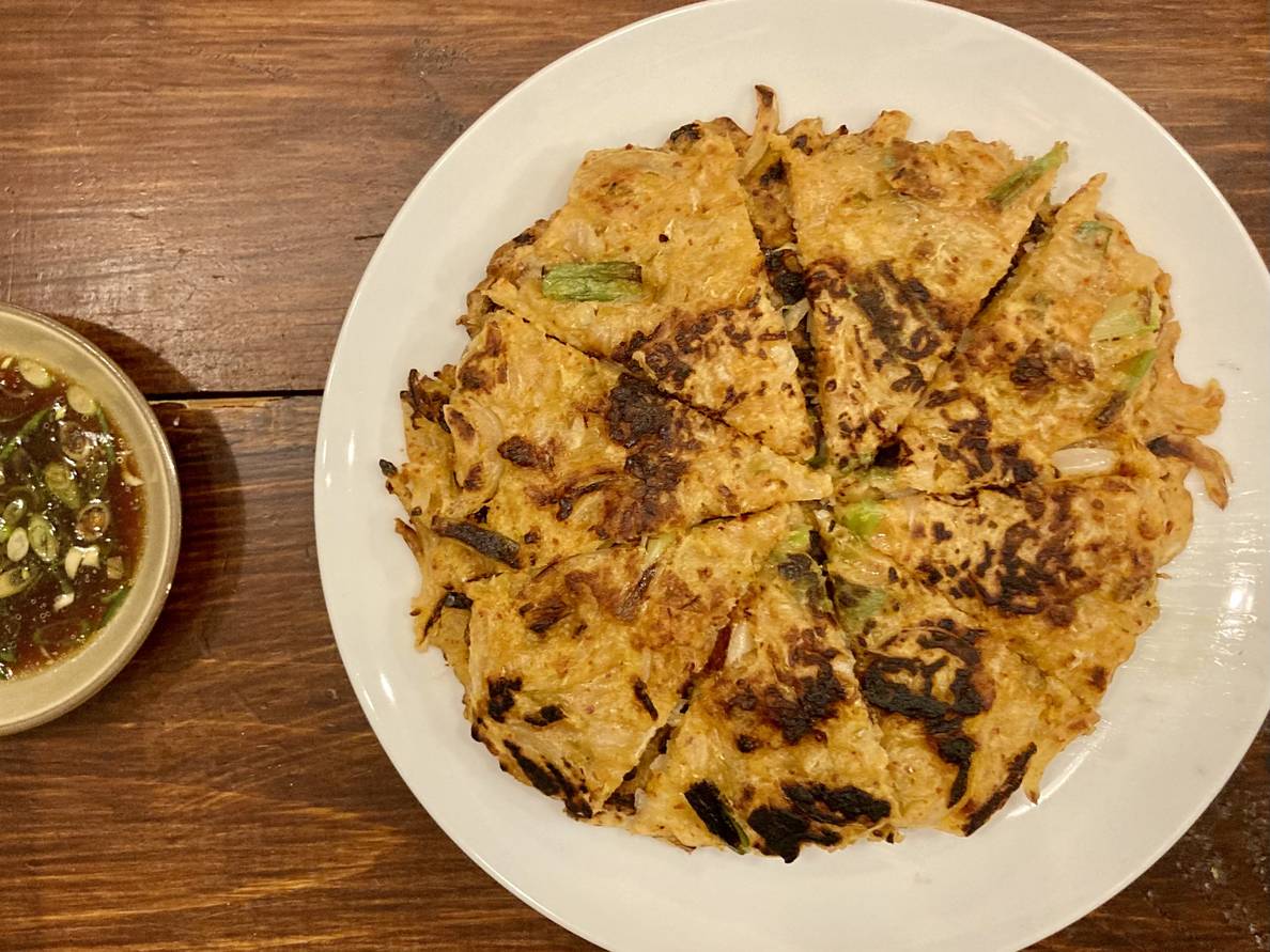 A photo of Korean kimchi pancakes on a plate on a table. The pancakes have been cut into eighths, and bits of scallions are visible. A bowl of dipping sauce is next to it.