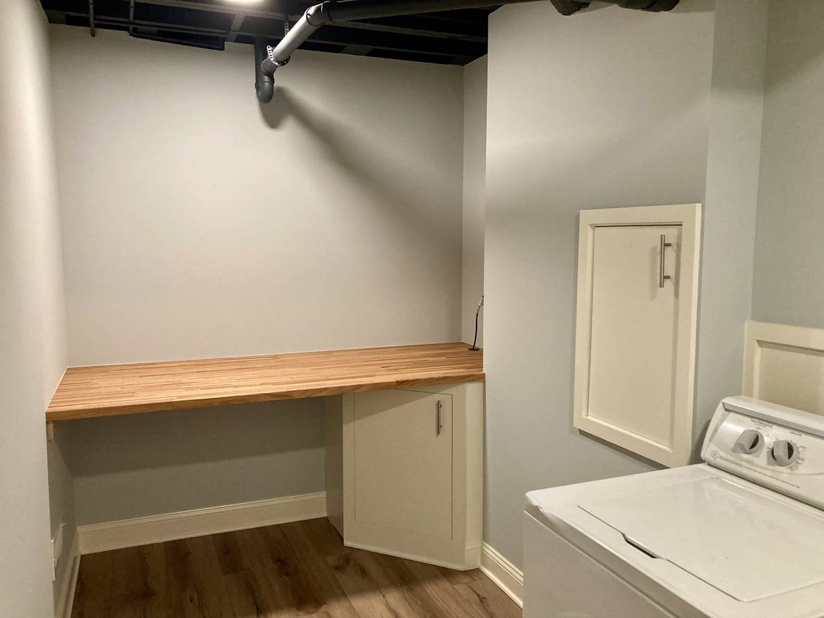 A photo of the finished laundry room from one of the corners. Part of the washing machine is visible from the right side, with some panels built into the wall behind it that provide access to plumbing. On the far wall is a butcher block shelf with a cabinet underneath that provides access to the sump pump.