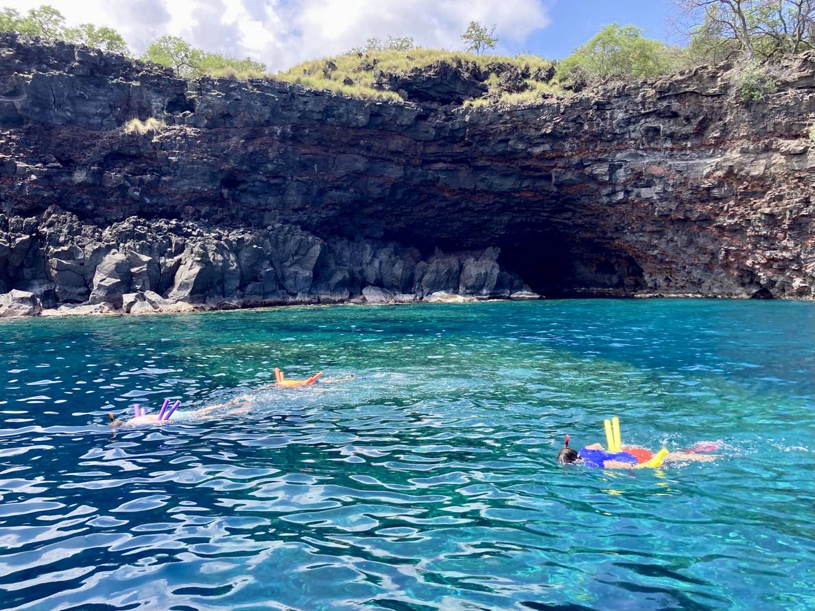 A photo of tropical water with a few snorkelers in it. A rocky cliff rises from the water in the background.