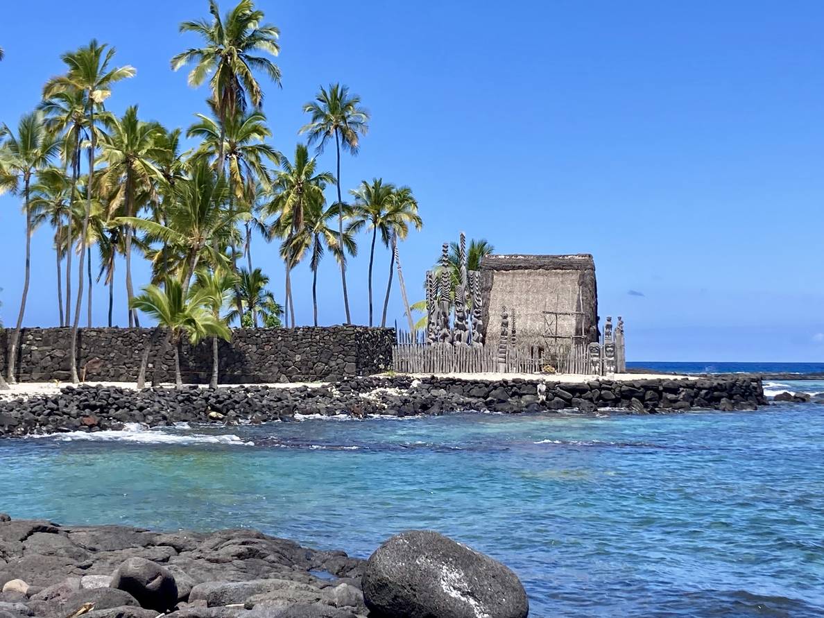 A photo of a thatched Hawaiian structure across a small bay. Behind the structure are several palm trees. The sky is cloudless and the water is unbelievably clear.