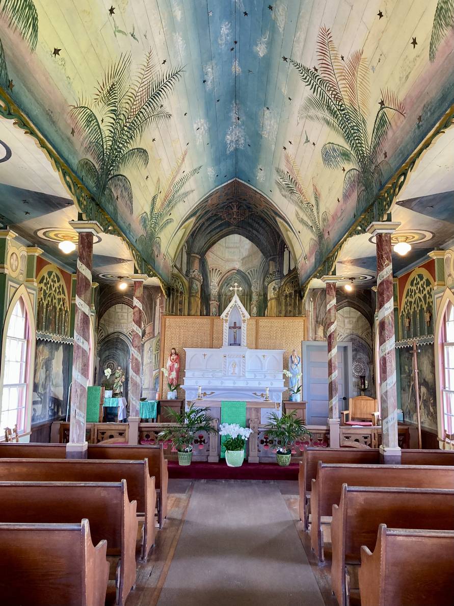A photo of the interior of a small church. Everything is painted in murals. The columns are topped with paintings of palm tree tops on the ceiling. The walls are a series of murals.
