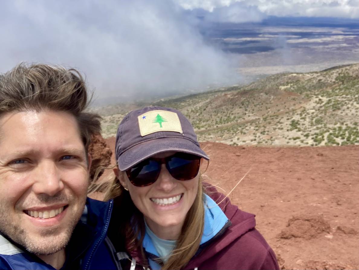 A photo of a wind-blown man and a woman. A cloud obscures half of the background. The other half, in the distance is scrubby. In the foreground the ground is scree.