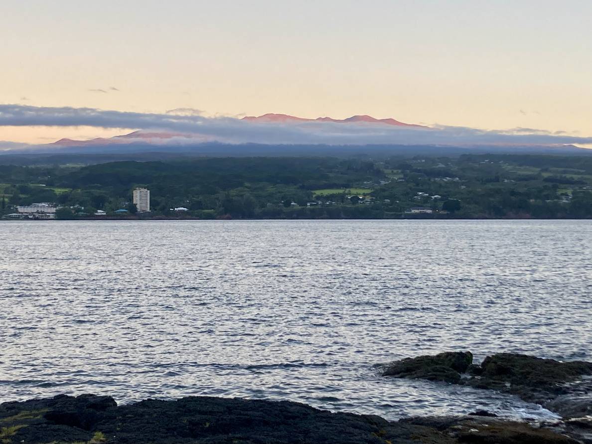 A photo of Mauna Kea, with a wisp of cloud, across the water. The sun is lighting the peak while everything else is still in the shade.