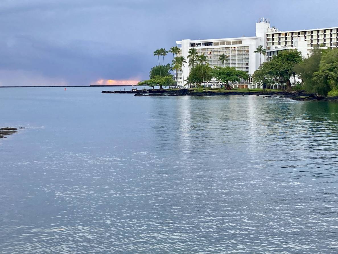 A photo of hotels on along the shore in Hilo. Palm trees line the shoreline. Sunrise light peeks through the clouds right at the horizon.