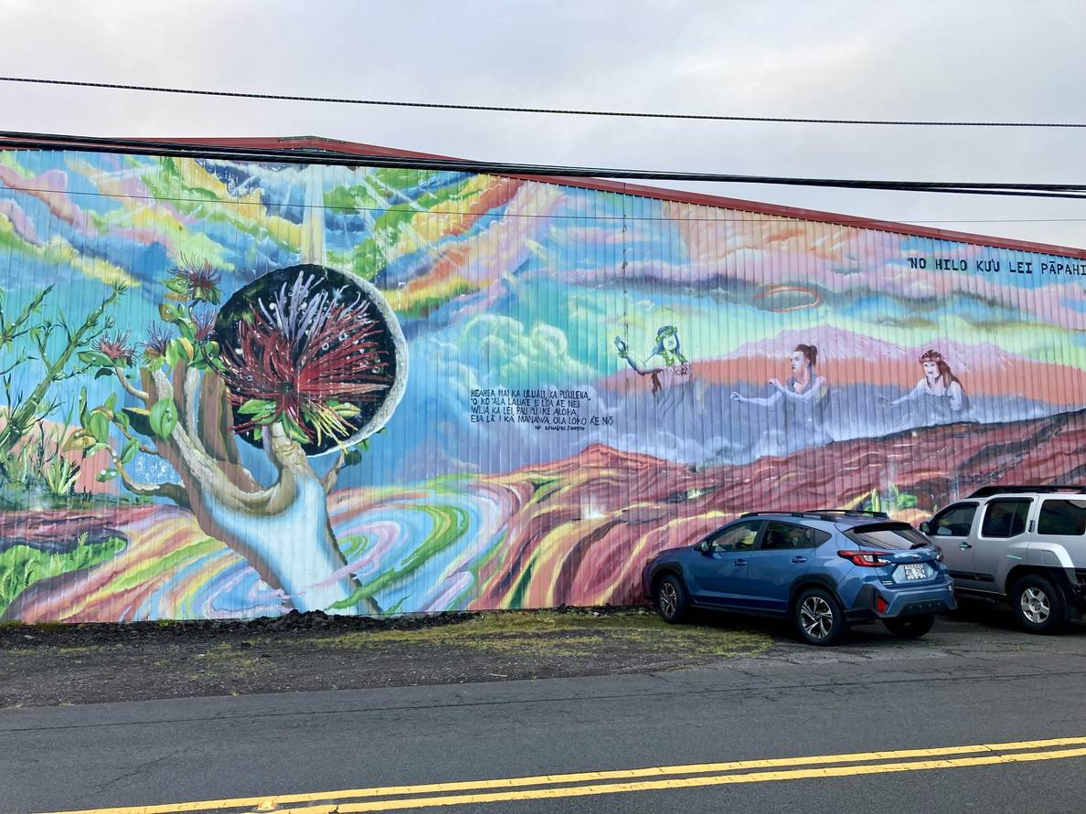 A photo of a mural on the side of a building. The painting has a hand holding an orb with a flower in it. An upside down rainbow is over that. Hawaiians rise out of the clouds in front of Mauna Kea on the right.