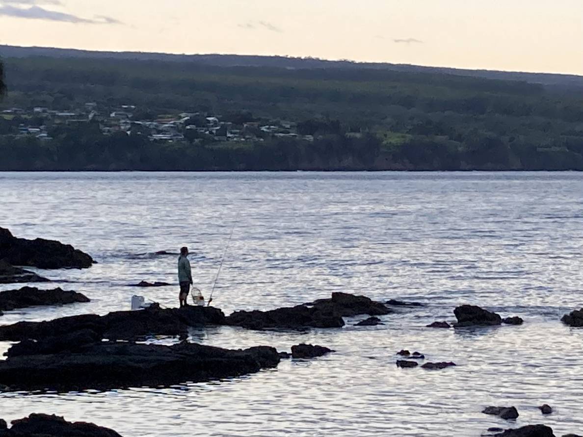 A photo of a fisherman on rocks in the water silhouetted against water and land on the other side of the water.