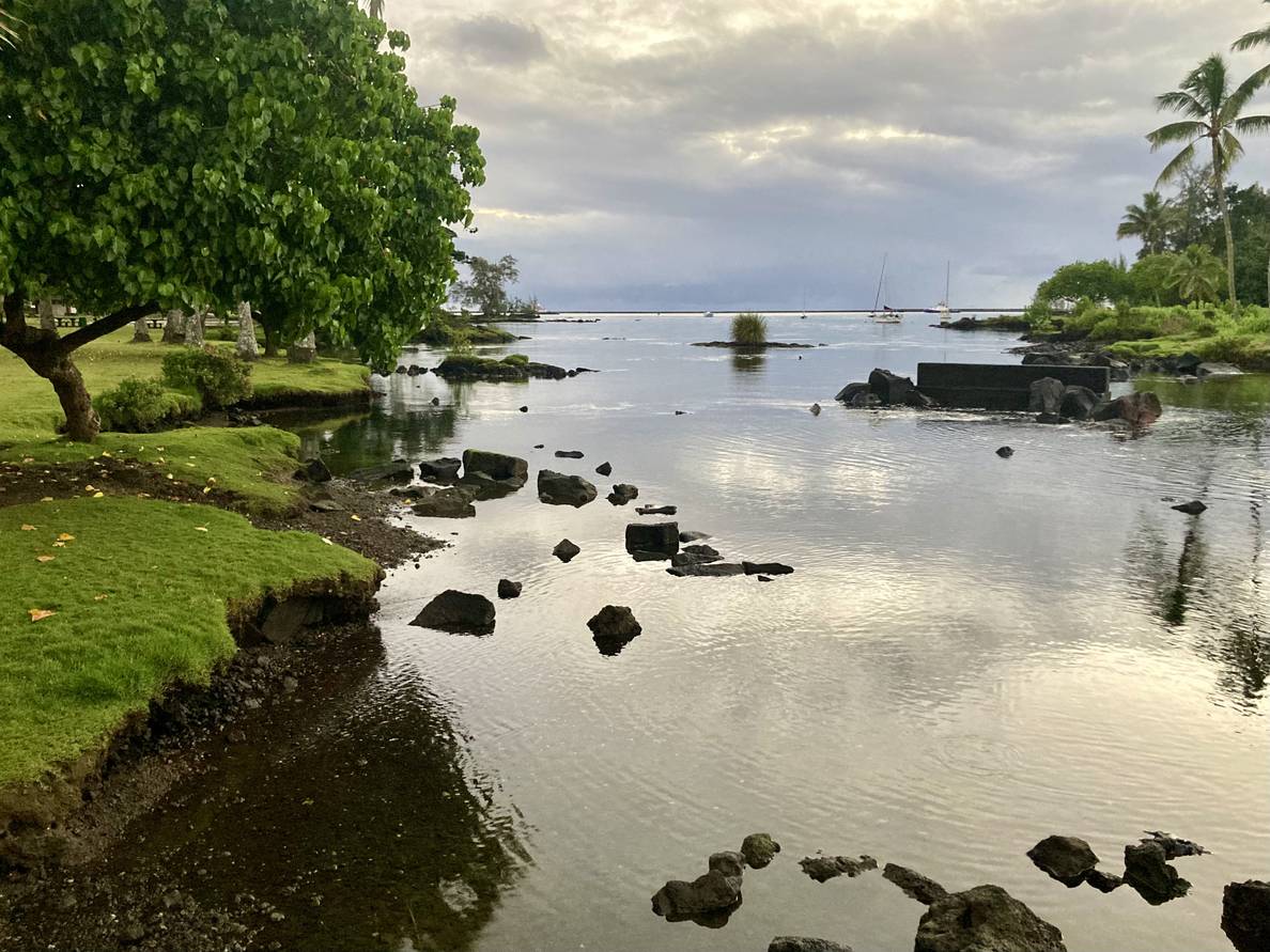 A photo of calm water flanked on both sides by a green shoreline. Rocks poke above the surface of the water. The sky is cloud with sunrise light peeking through.