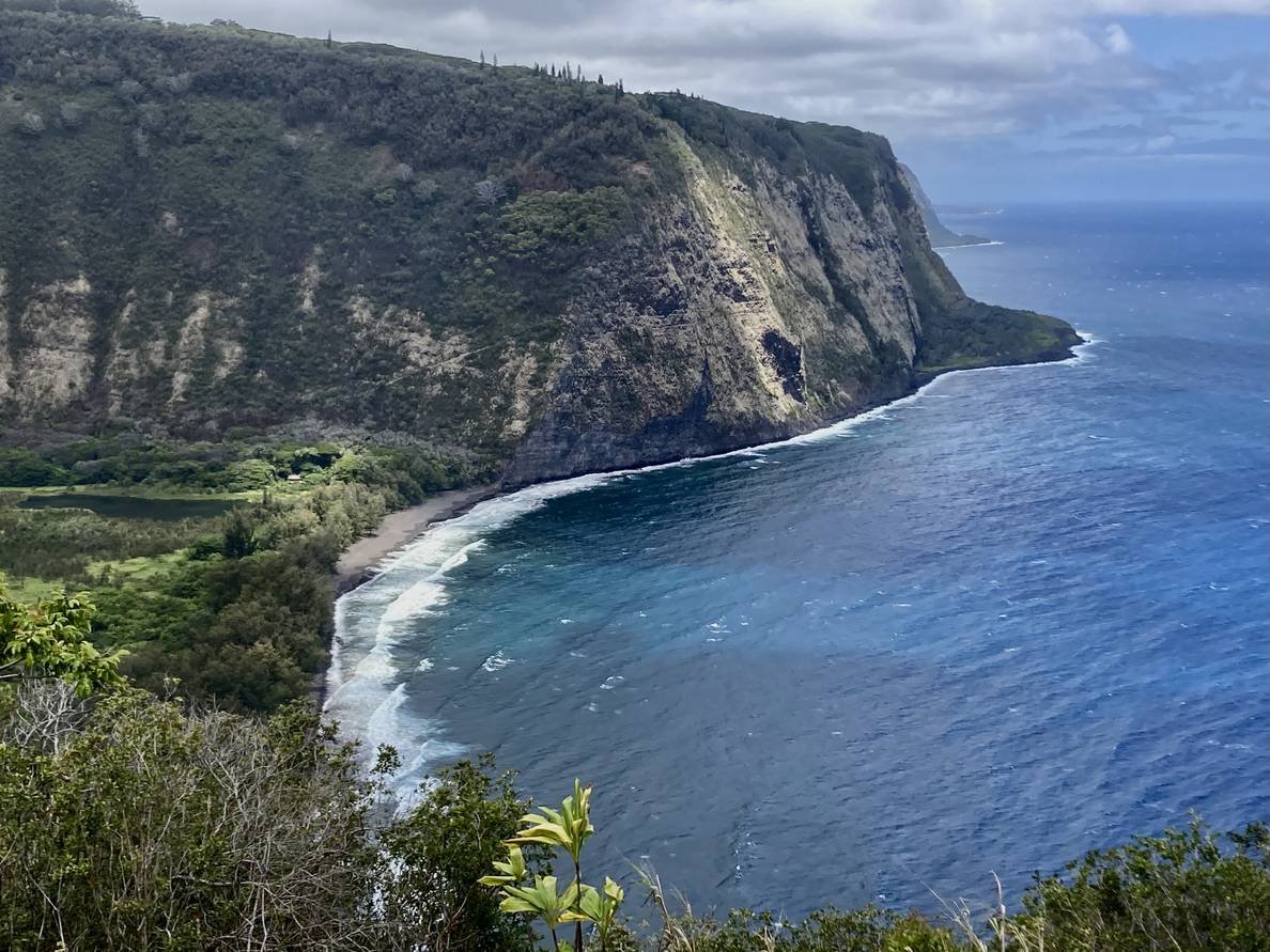 A photo of a valley meeting the ocean. The valley is lush and lined by steep cliffs. The beach is black sand.