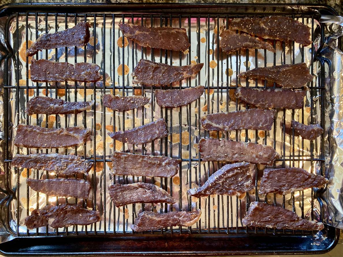 A photo of beef slices on a wire rack in a sheet pan. The meat has been cooked and some drippings can be seen cooked onto the sheet pan.