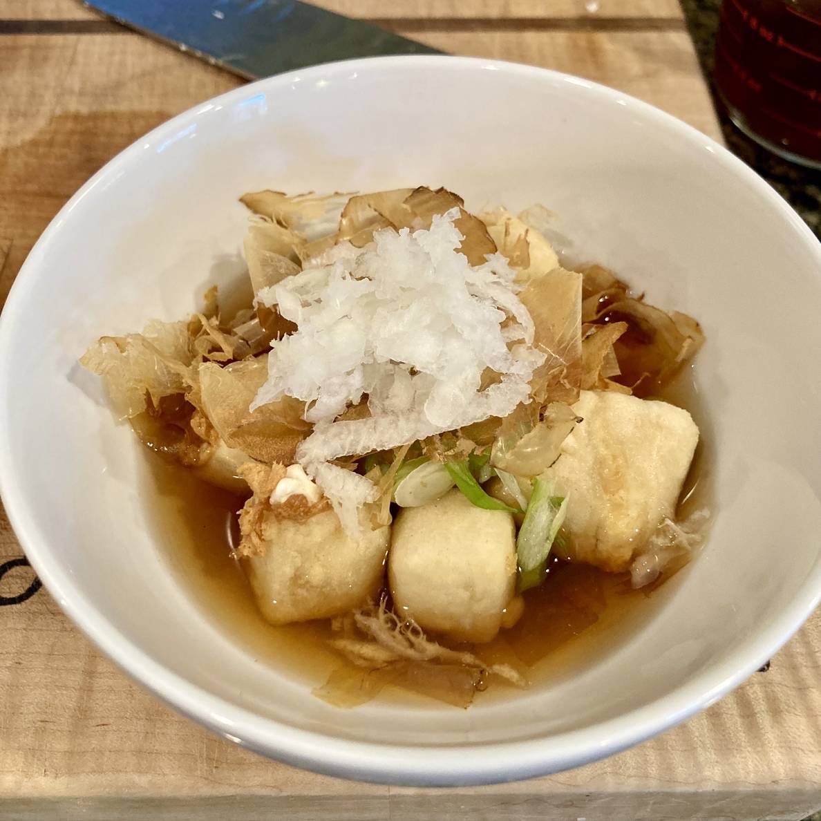 A photo of a bowl of fried cubed tofu in a soy dashi sauce with green onions, katusobushi, and grated daikon radish on top.