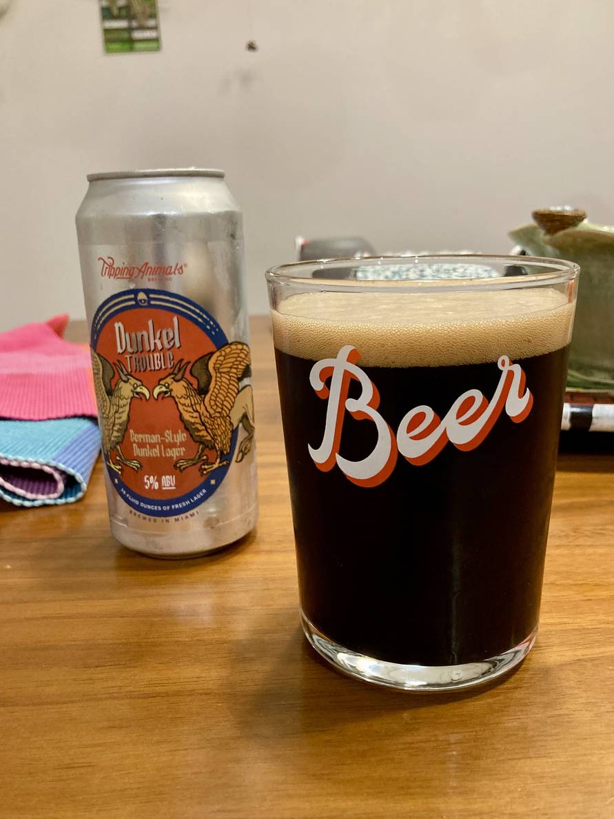 A photo of a glass of dark beer with a can behind it on a table. The glass has the word “Beer” printed on it. The can is a tall boy with a picture of two griffins facing each other. It’s a “German-Style Dunkel Lager.”