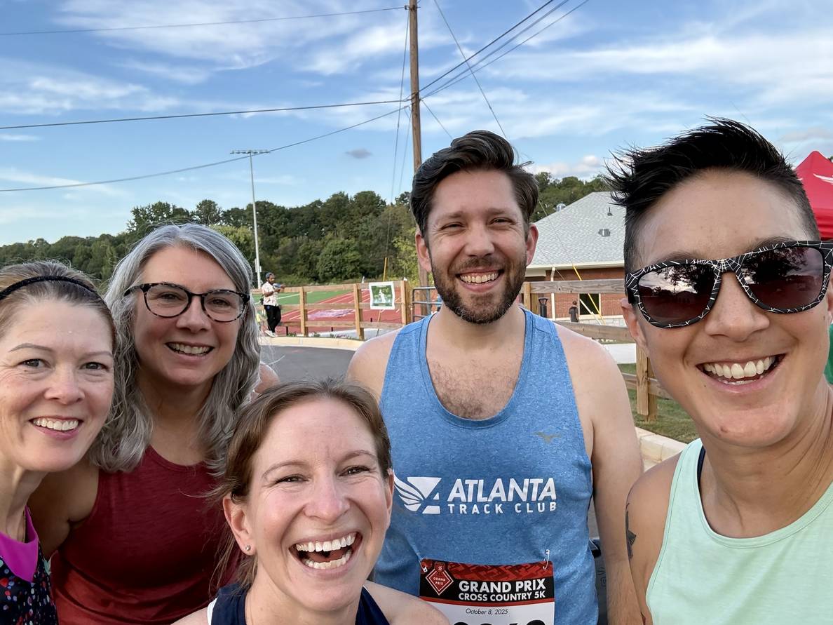 A photo five people, most in running attire, laughing and smiling at the camera. Behind them is a running track, public restrooms, and trees beyond that.