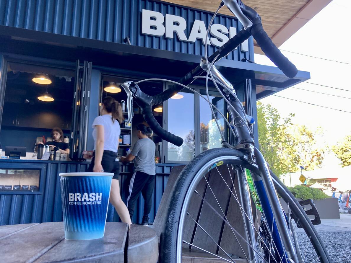 A photo of the Brash coffee kiosk on the BeltLine. A bicycle is leaned against a chair and a paper cup that says “BRASH COFFEE ROASTERS” is on a small table.