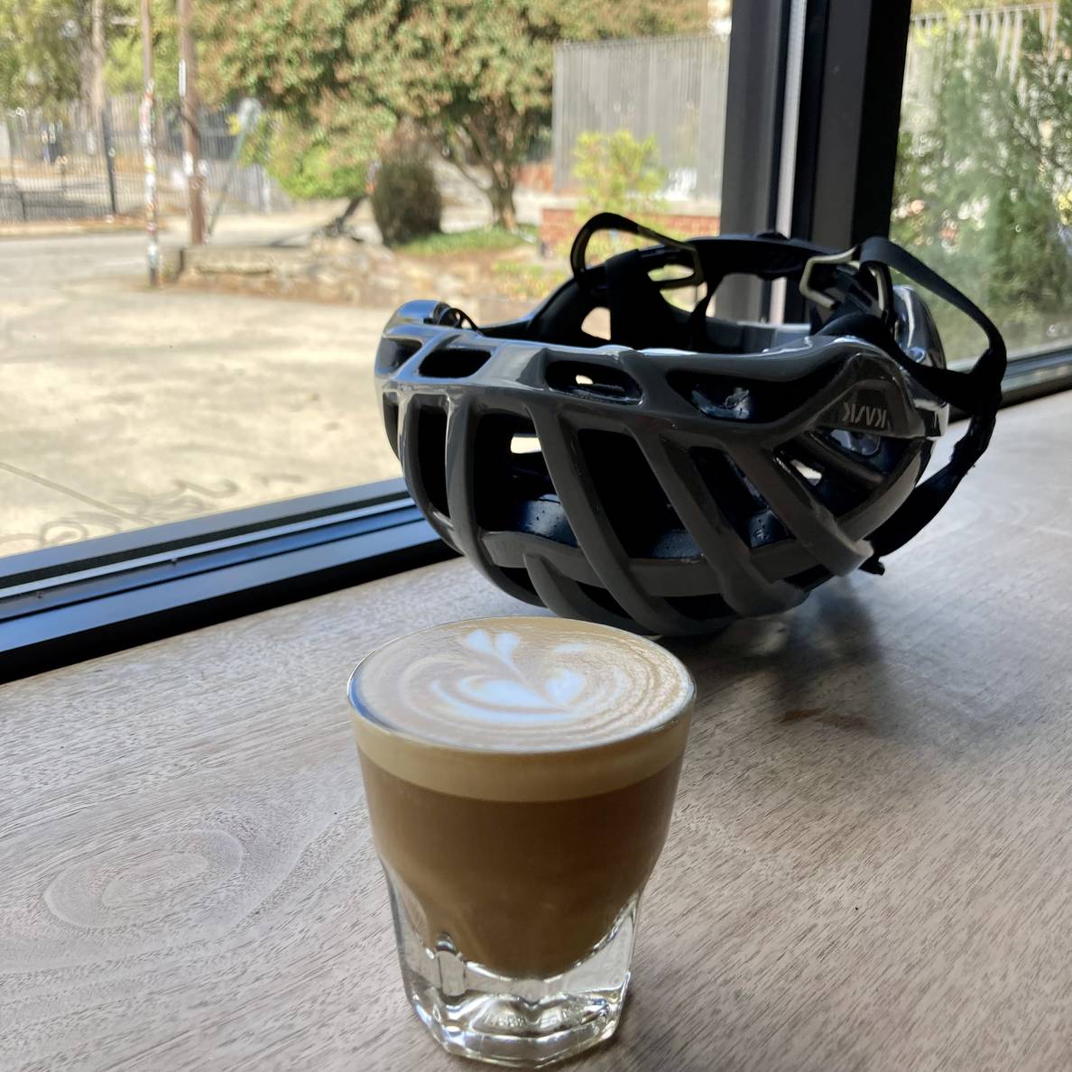 A photo of a cortado in a glass cup on a wooden counter. A bike helmet sits upside down behind it. Windows look out onto a small parking lot and smaller patio space.