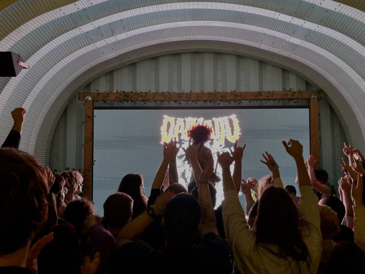 A photo of a band on a low stage. A guitarist stands on the base drum with his back to the crowd. The silhouetted crowd is pressed close to and on the stage with hands in the air.