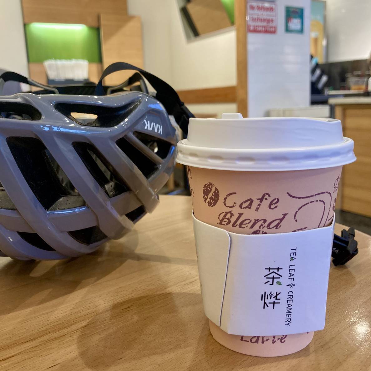A photo of a to-go cup on a table with a bicycle helmet behind it. The coffee sleeve says “Tea Leaf & Creamery.”