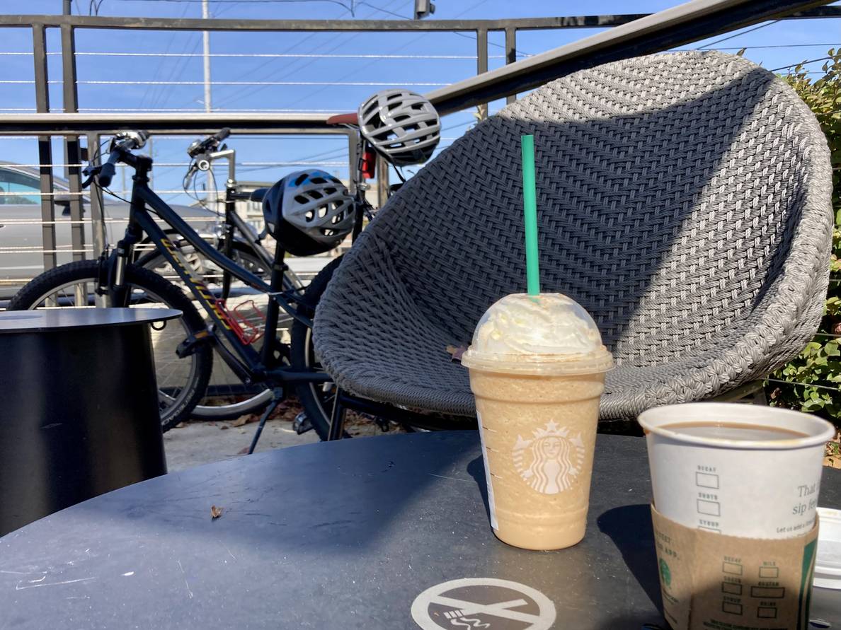 A photo of a black coffee and some kind of Frappuccino on an outdoor table. Two bicycles are in the background.