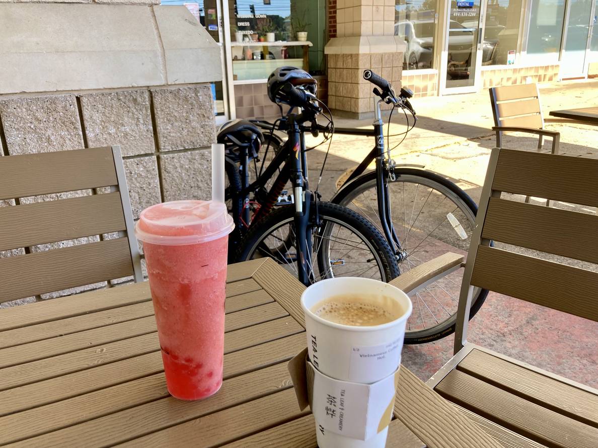 A photo of a coffee and a strawberry bubble tea on an outdoor table with two bicycles behind it.