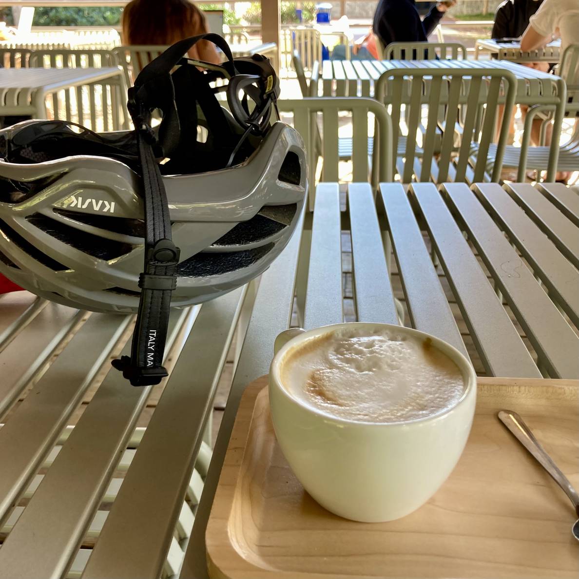 A photo of a wooden tray with a cappuccino on an outdoor table. Behind the tray is a bicycle helmet.