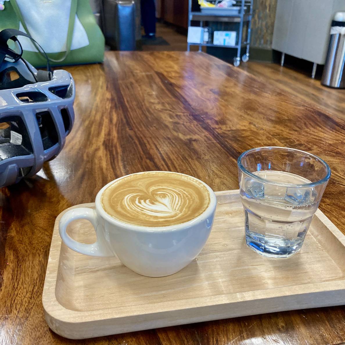 A photo of a cappuccino in a 6oz mug with a small glass of seltzer on a wooden tray on a wooden table. Peeking into the frame is my bicycle helmet.