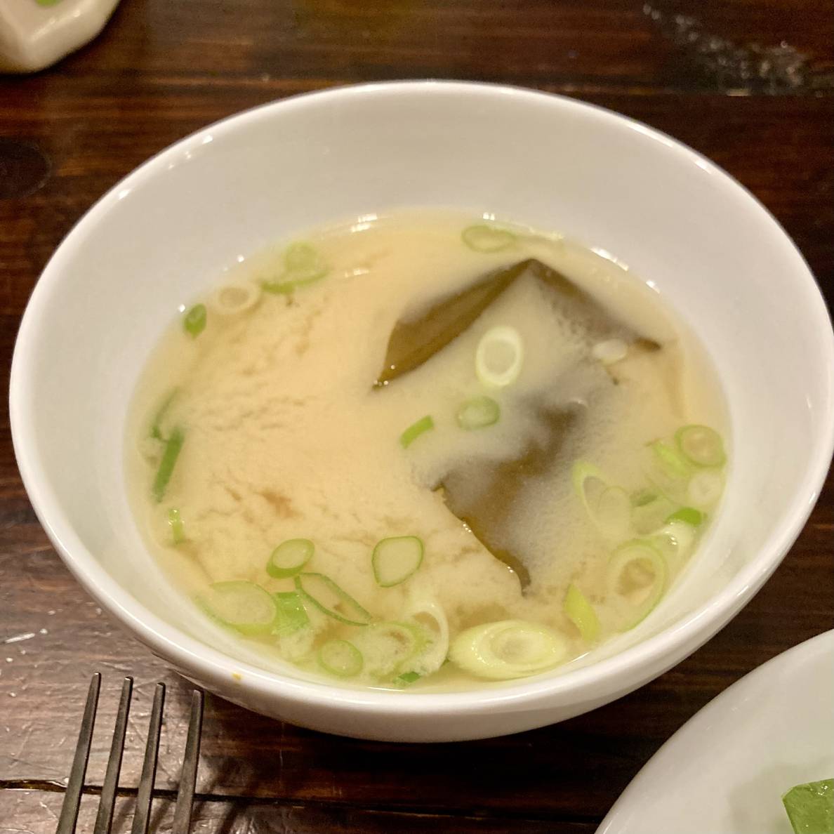A photo of a bowl of the finished miso soup. It is quite cloud from the miso. A large piece of alaria “wakame” is visible, and several scallions float on top.