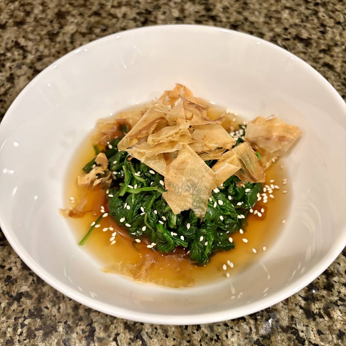 A photo of the final dish. The spinach sits in a mound in the middle of a bowl with the sauce around it. Sesame seeds are scattered on top and then large flakes of katsuobushi.