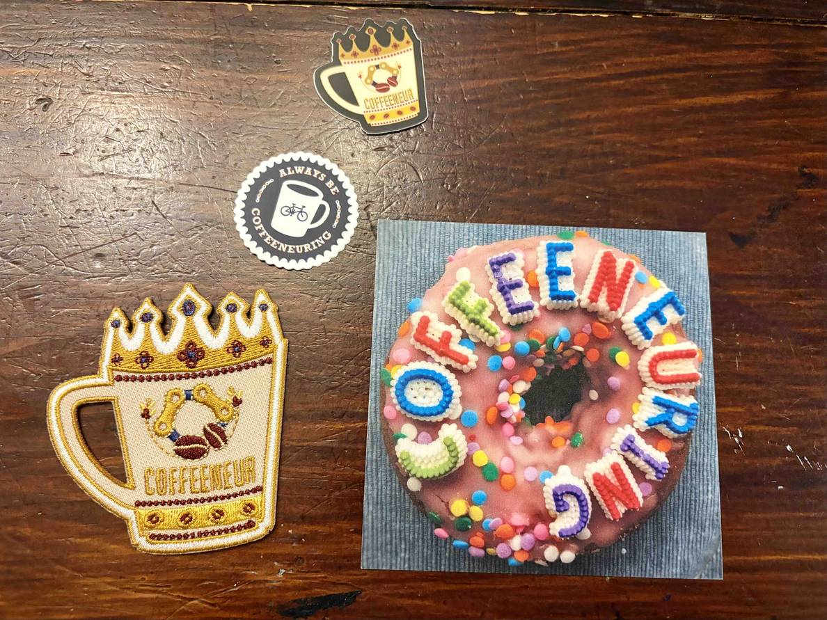A photo of stickers, a patch, and a small print, all themed for this year’s coffeneuring challenge.