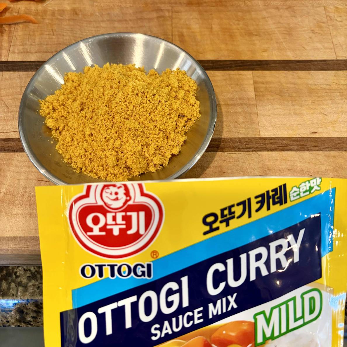 A photo of a 3 tablespoons of the curry powder in a small dish with the package held in front of it. The package reads “Ottogi Curry Sauce Mix Mild”