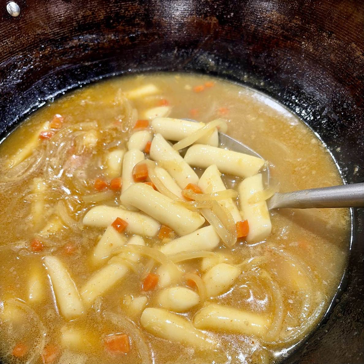 A photo of the stew, mid-simmer, in the wok. Some of the rice cakes are more visible from a spatula holding them from the broth.