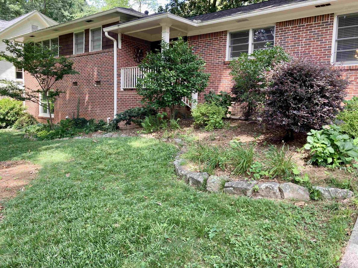 A photo of the front entry area. Steps from the front door go along the front of the house, hidden by large shrubs. The bed the shrubs are in curves out from the house with some additional perennials and a rock border. A narrow swath of grass is in front of that bed, with the driveway to the right.