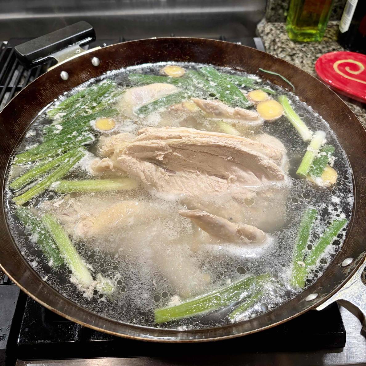 A photo of the stock cooking in a wok. The chicken carcass pokes out of the surface. A lot of green onions float on the surface along with some ginger slices.