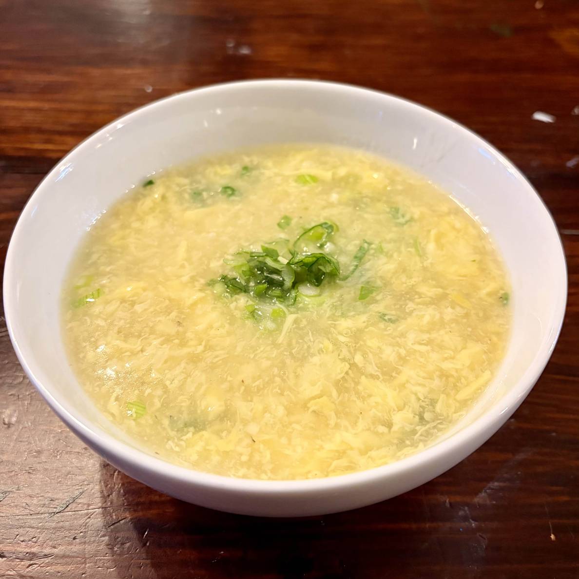 A photo of a bowl of the egg drop soup. It is intensely eggy with a some scallions in the soup but a larger pinch in the middle.