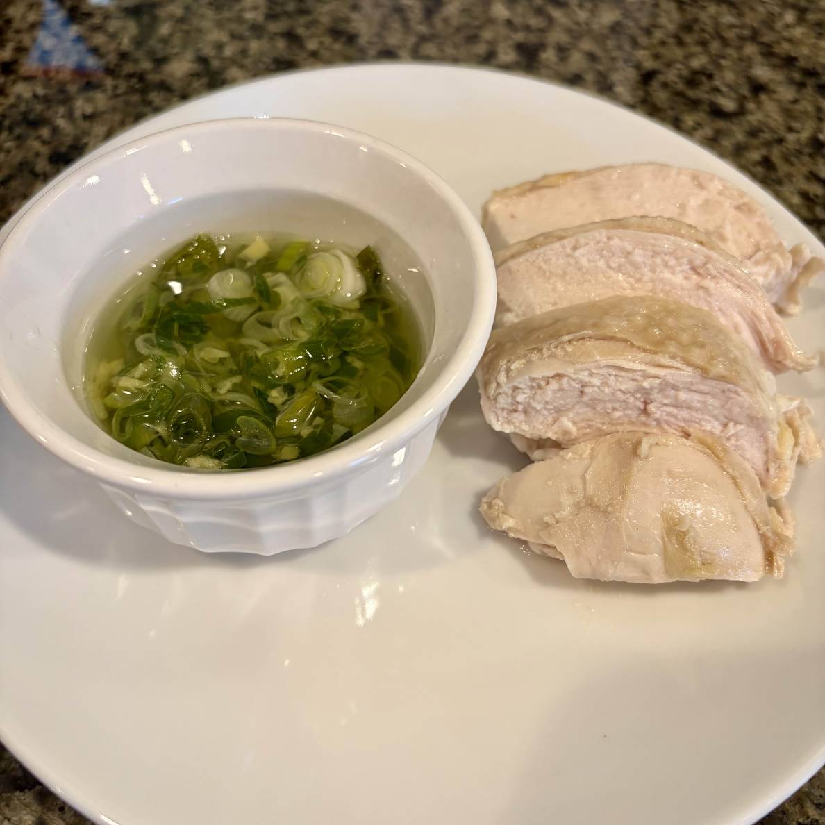 A photo of a few slices of the poached chicken on a plate with a ramekin of the fragrant scallion-ginger oil.